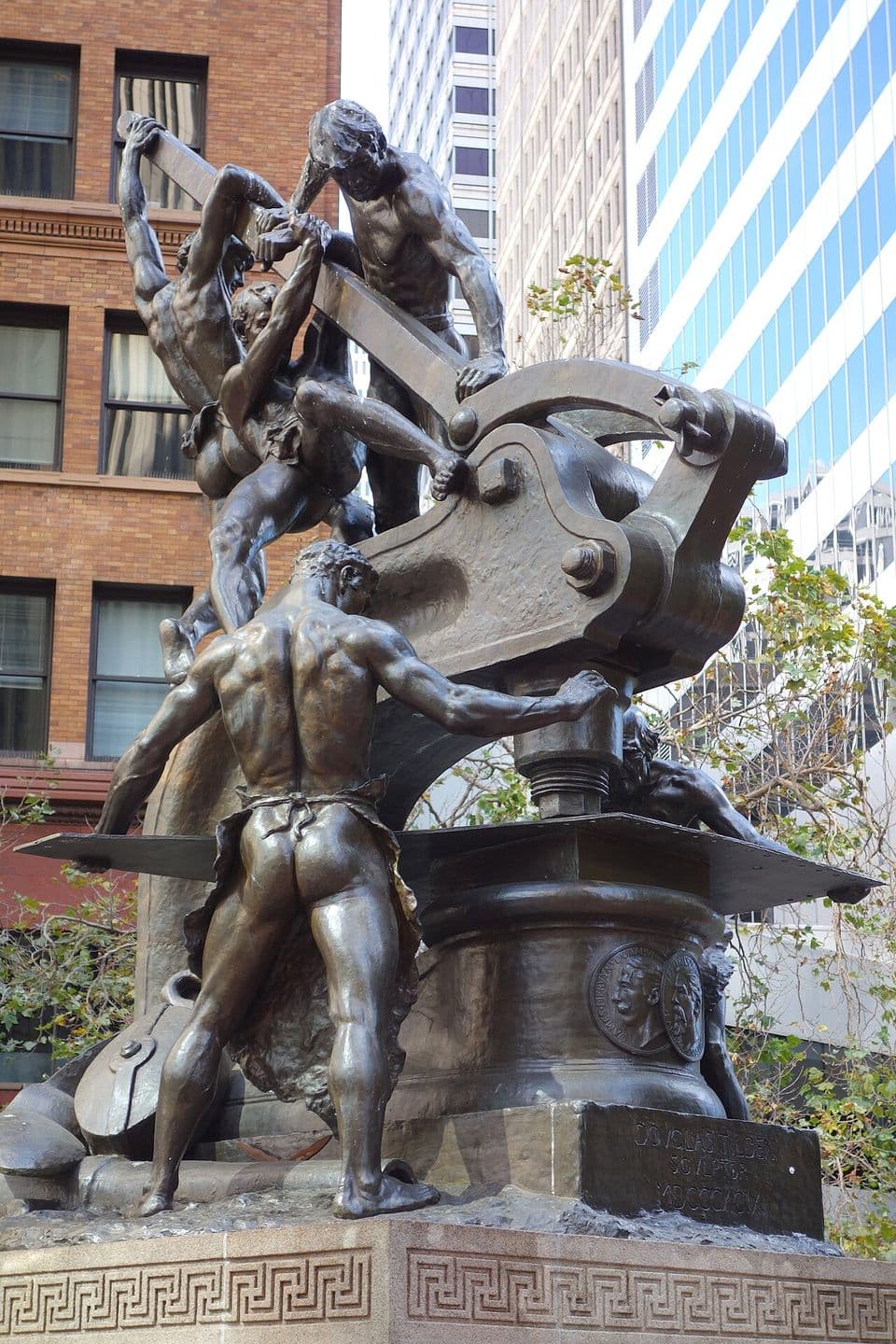 Douglas Tilden's bronze Mechanics Monument: muscular workers straining at a giant punch press, on a granite base carved with a Greek key, with downtown towers behind.